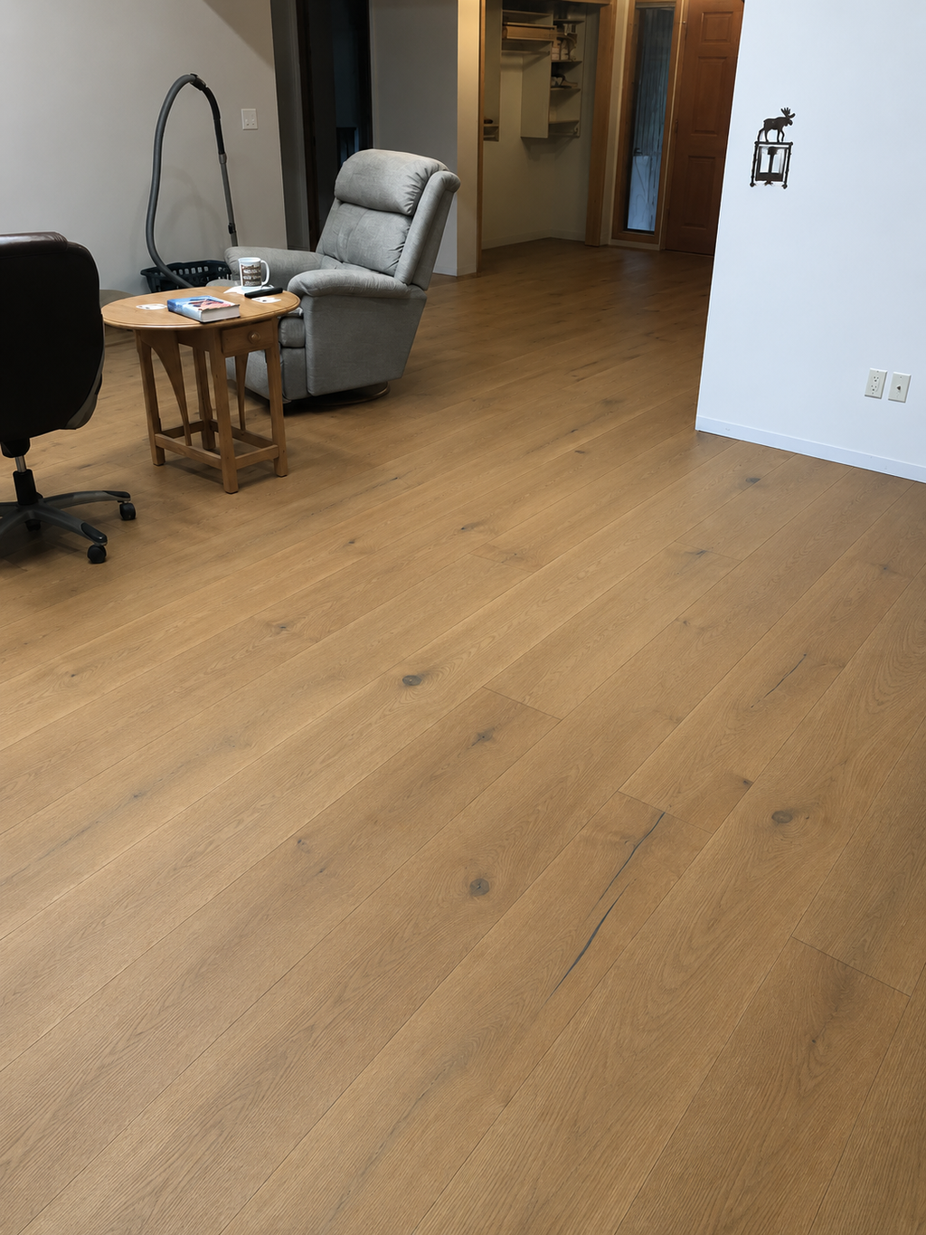 Light brown wood-look laminate or vinyl plank flooring shown in a home office/living room setting with a gray recliner, wooden side table, and office chair visible.