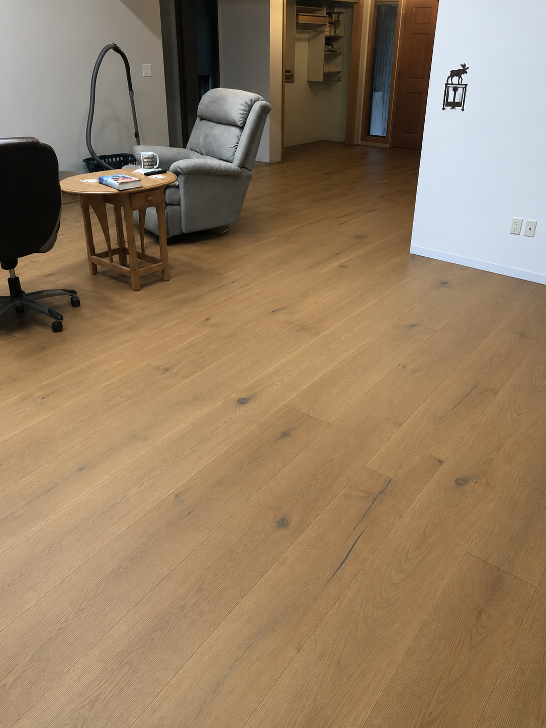 Light brown wood-look laminate or vinyl plank flooring shown in a home office/living room setting with a gray recliner, wooden side table, and office chair visible.